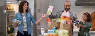 family unpacking their groceries. father is passing the mother a milk in paper carton to put in the fridge.